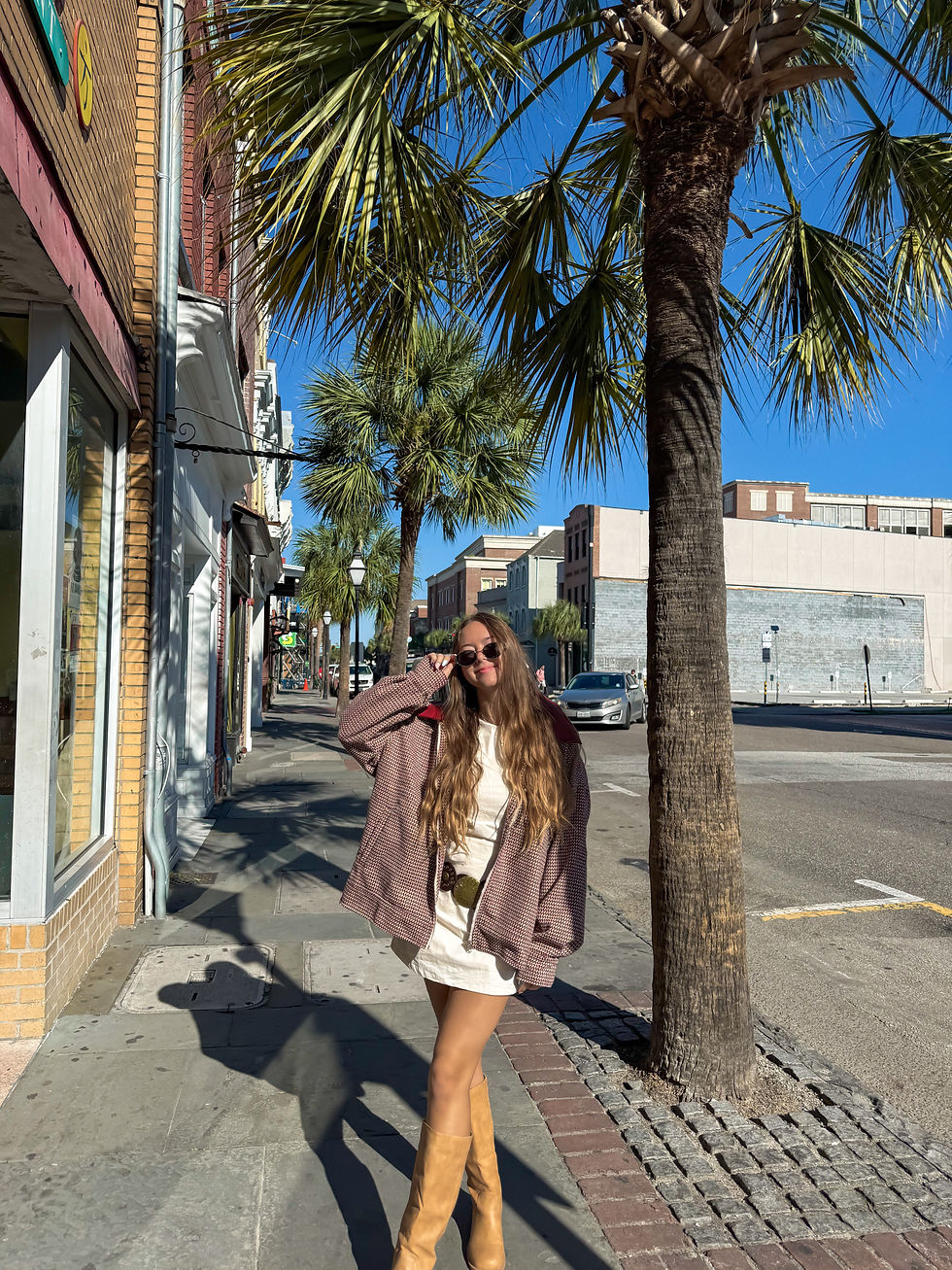 Woman with long hair in sunglasses and a brown jacket poses on a sunny street lined with palm trees. Blue sky and buildings in the background.