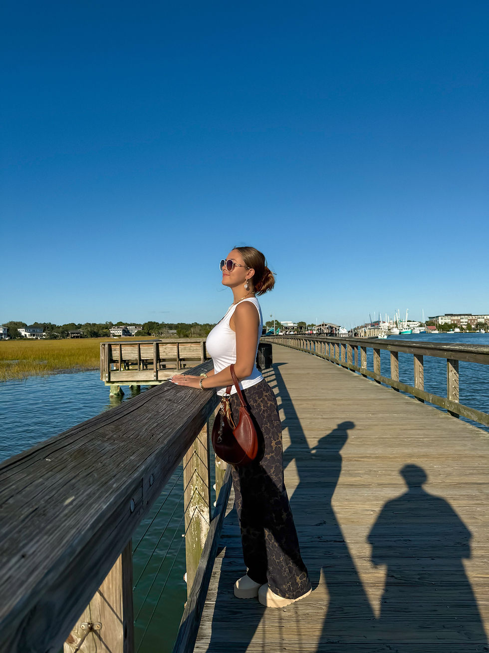 Woman in white top and sunglasses stands on wooden pier, gazing at water. She holds a brown bag. Clear blue sky, bright day.