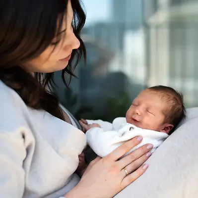 Lifestylové newborn focení miminek Ostrava, doma: Maminka drží usmívající se a spící miminko na kolenou během lifestyle focení na balkoně v Ostravě. Přirozená rodinná fotografie v domácím prostředí.