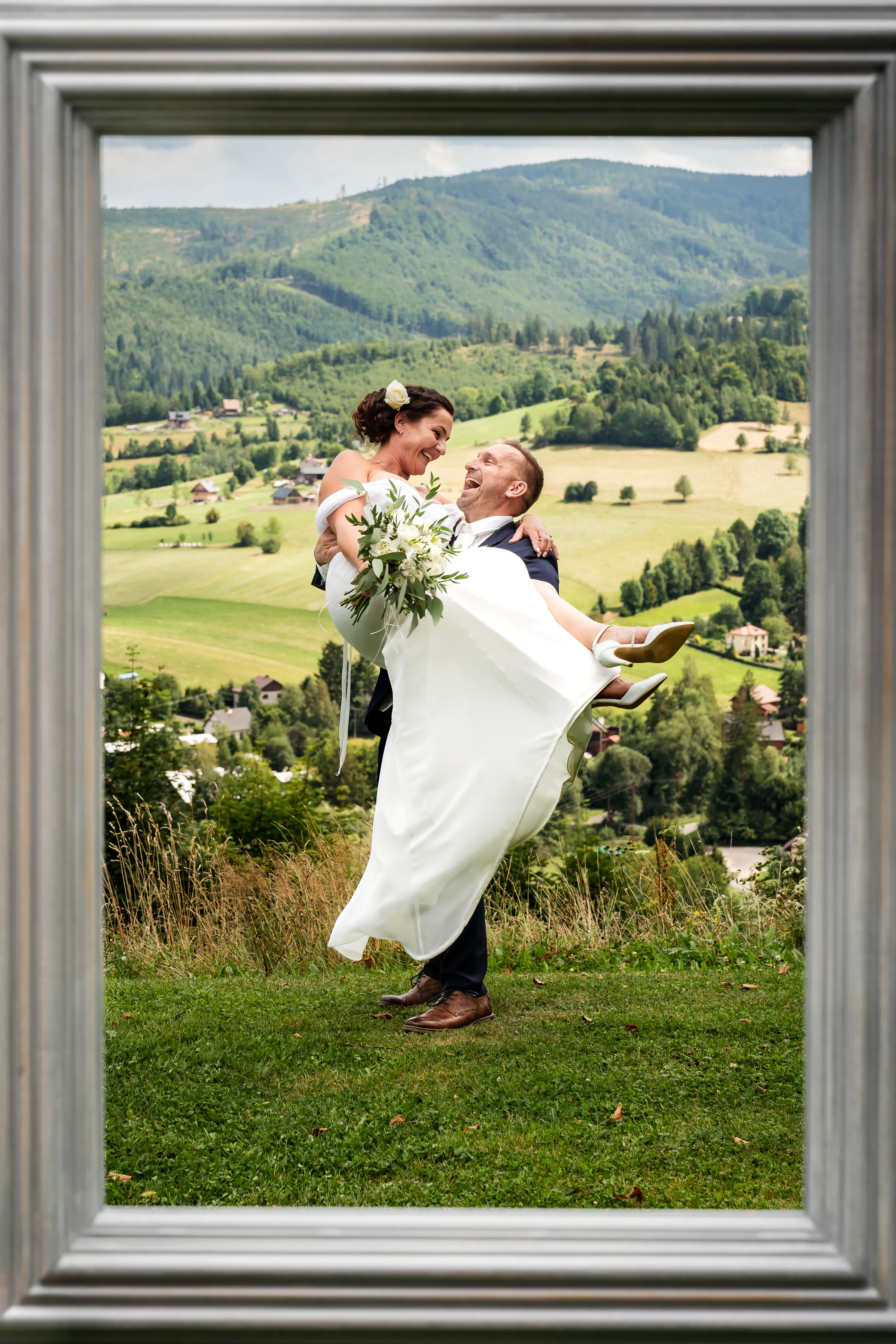 Romantická svatební fotografie, na které ženich drží nevěstu v náručí, zatímco se na sebe zamilovaně smějí. Kompozici doplňuje rám obrazu v popředí, který dokonale orámuje scénu s panoramatickým výhledem na hory v pozadí. Svatební fotograf Ostrava, Moravskoslezský kraj. Svatební fotky s atmosférou. Svatební fotografka. Originální svatební fotky.