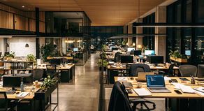Escritório moderno vazio à noite, com mesas, computadores, plantas e quadro branco sob luzes quentes.