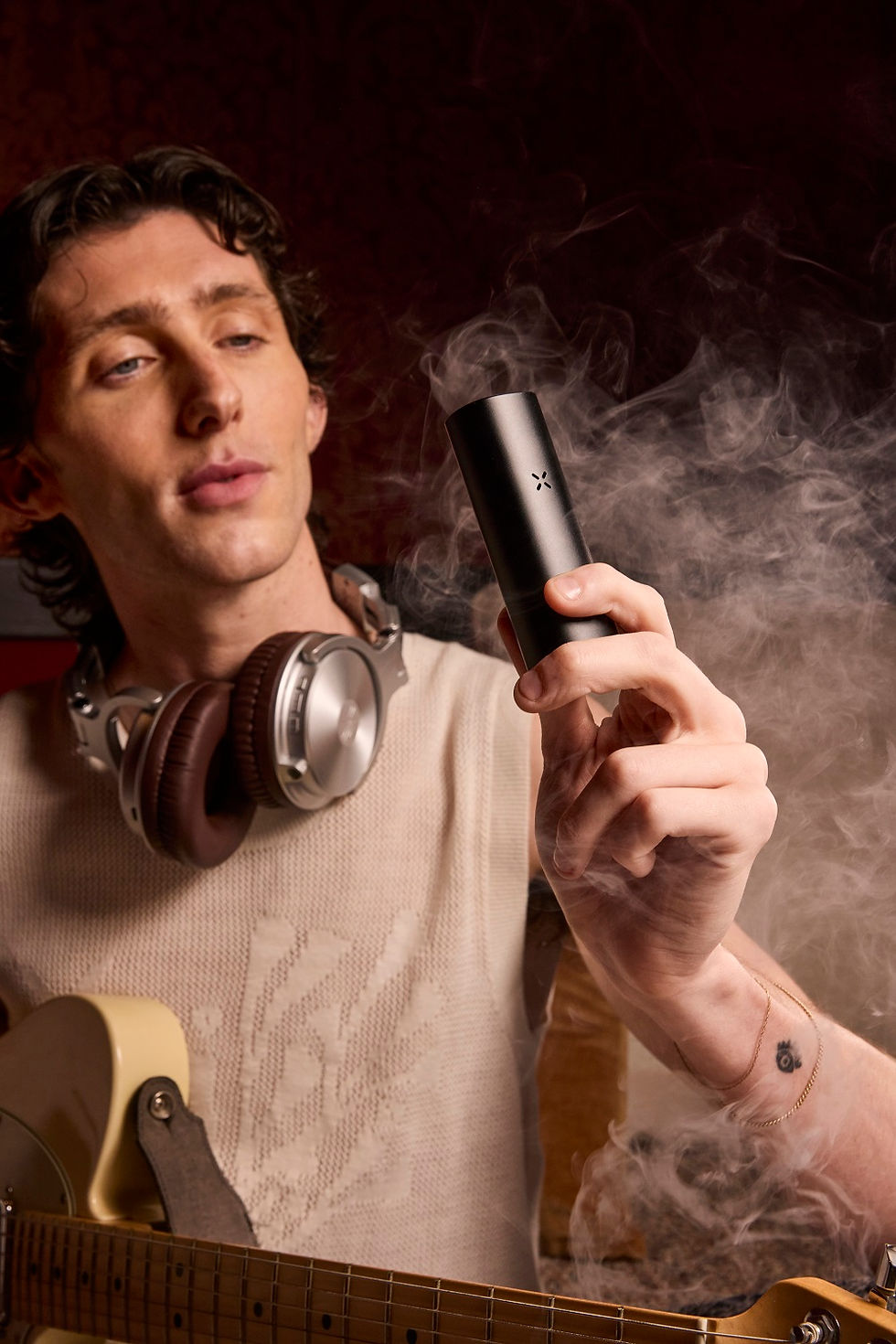 A person with headphones and a guitar holds a black PAX vaporizer releasing vapor in a warm, creative setting.