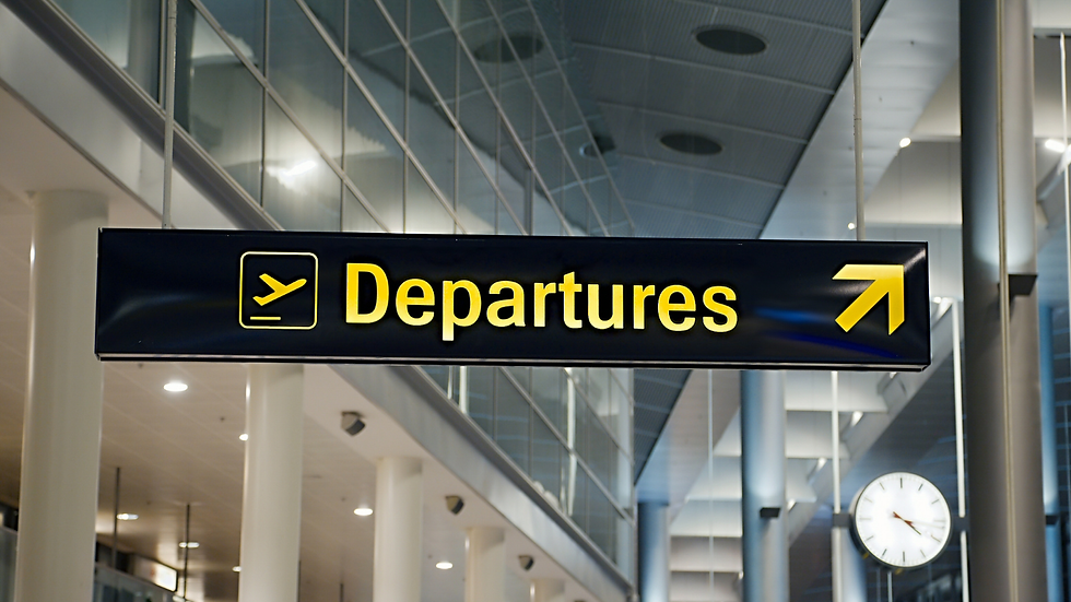 Airport sign showing "Departures" in yellow with an arrow, hanging in a modern terminal. Ceiling panels and a round clock are visible.