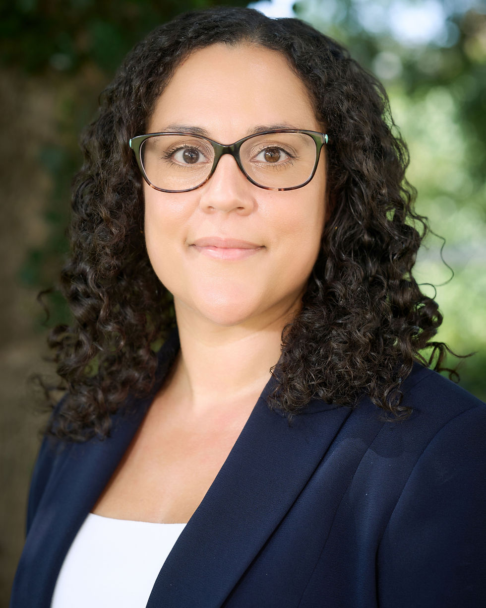 Portrait of a woman with curly hair and glasses in a navy blazer, smiling softly against a blurred green outdoor background.