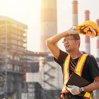 Hot construction worker in safety vest removes a yellow hard hat at an industrial plant, holding a clipboard in bright sunlight.