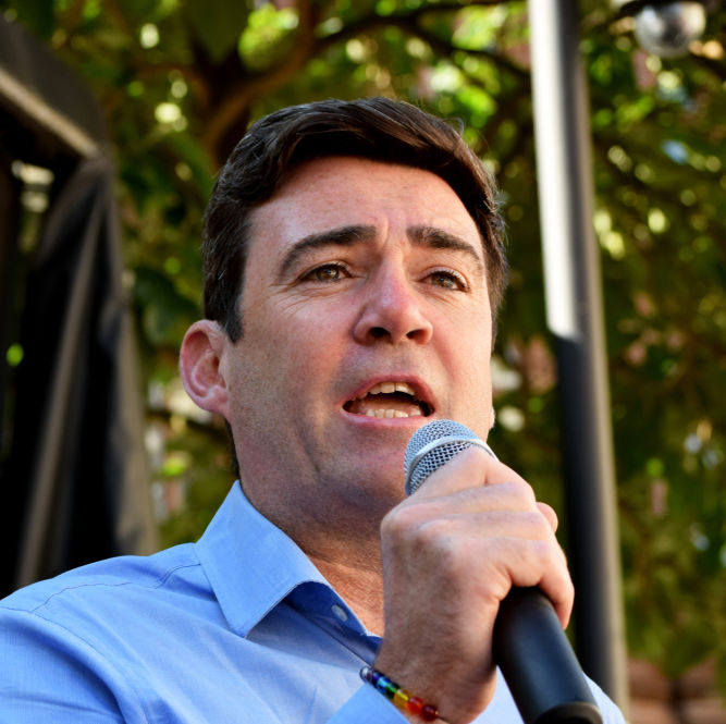 Man in a blue shirt speaks into a microphone outdoors, with blurred green trees in the background.