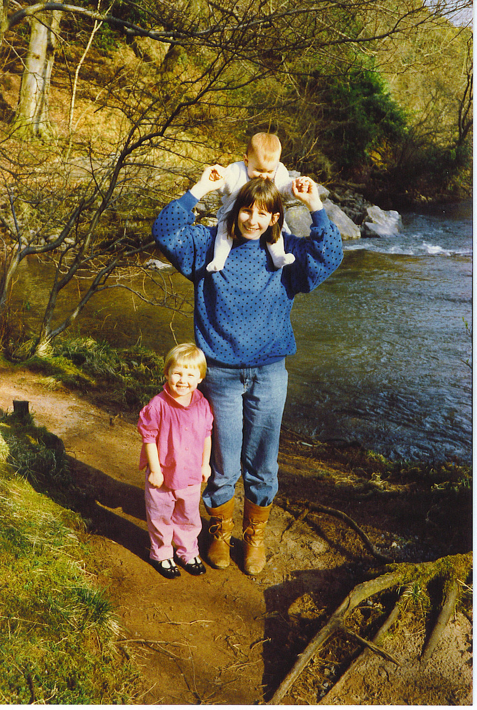 A Mum stands next to a river with a child next to her and a baby on her shoulders
