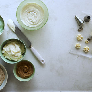 Bowls of pastel-colored frosting, piping bag, and nozzles on a white surface, with star-shaped decorations being piped.