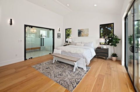 Modern bedroom with a white bed, wooden bench, and black-accented nightstand. Large windows and sliding glass doors.