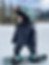 A toddler in a puffy navy snowsuit and teal helmet stands on a snowboard in a snowy area, looking to the side. Mountains and trees in background.