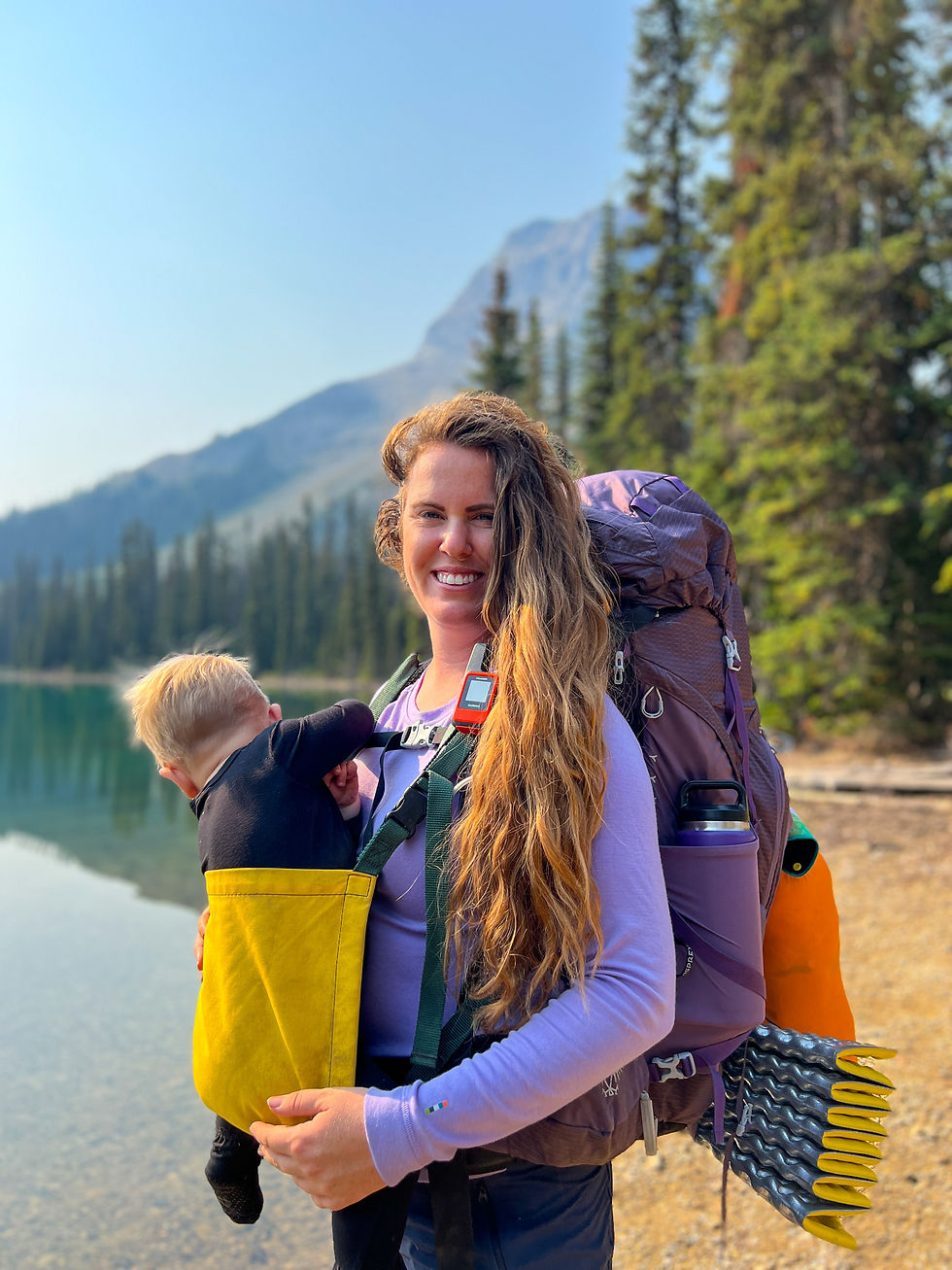 A woman with a backpack carries a baby in a yellow carrier by a lake, surrounded by forest and mountains. She's smiling under a clear sky.