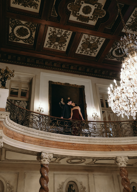 An engaged couple sharing a kiss on an ornate wrought-iron balcony overlooking a grand room with twisted marble columns, a large portrait painting, and a sparkling crystal chandelier.