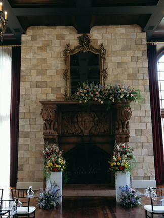 A grand indoor wedding ceremony space featuring a massive, intricately carved fireplace with lion motifs, set against a stone wall and topped with a large antique mirror. Lush, colorful floral arrangements decorate the mantle and two white pedestals at the base.