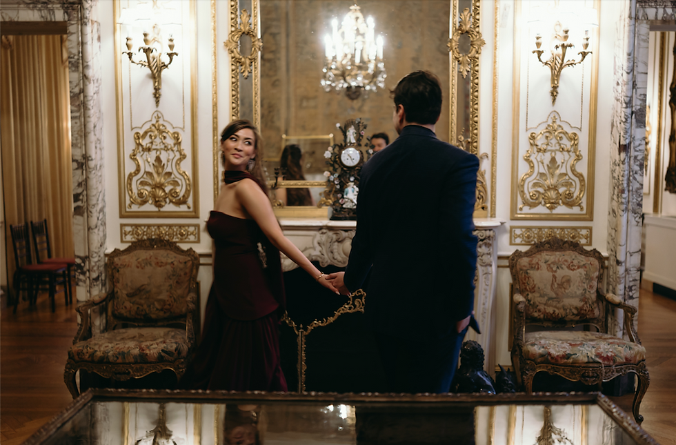 An engaged couple holding hands in a lavish room with gilded mirrors, a marble fireplace, and antique chairs. The woman is wearing an elegant burgundy gown and looking back over her shoulder at the man in a dark suit.