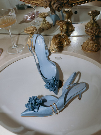 A pair of light blue satin slingback bridal heels with floral toe details resting inside a vintage white porcelain sink with ornate gold fixtures. A champagne coupe sits on the marble counter in the background.
