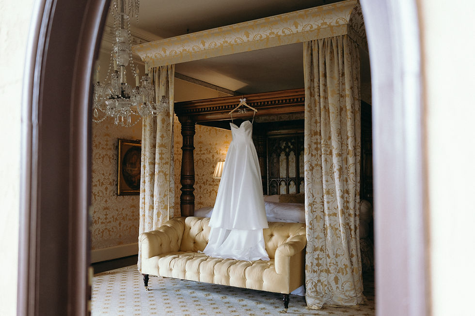 A classic white strapless wedding dress hanging from the heavy wooden canopy of a grand four-poster bed. The gown drapes elegantly over a tufted gold sofa in a luxurious bedroom featuring yellow damask wallpaper and a crystal chandelier, with the shot framed softly through a dark doorway arch.
