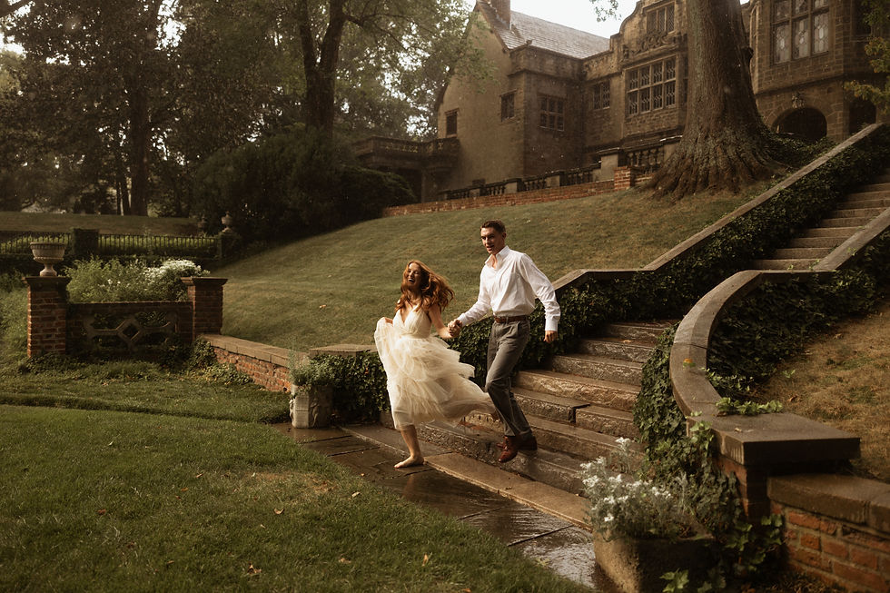 A joyful engaged couple running down sweeping, ivy-lined curved stone stairs in the pouring rain, with a grand historic stone mansion in the background. The woman is barefoot and wearing a layered white tulle dress.