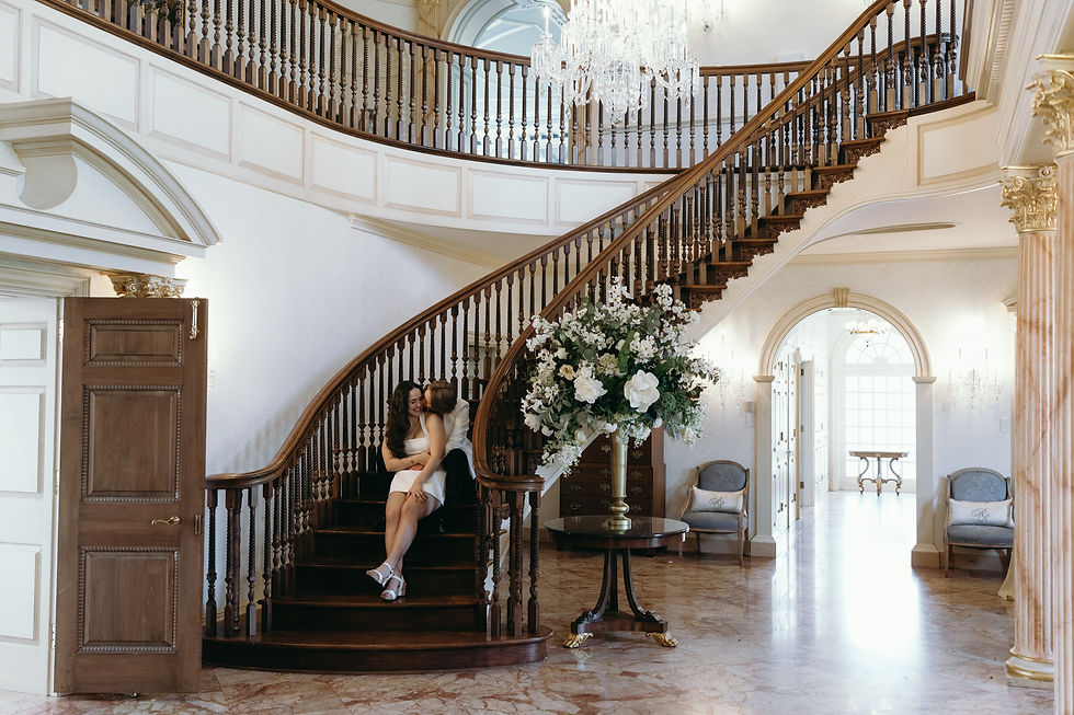 An engaged couple sharing a sweet moment sitting on the bottom steps of a grand curved wooden staircase at The Estate at River Run, with a large, elegant floral arrangement in the foreground on polished marble floors.