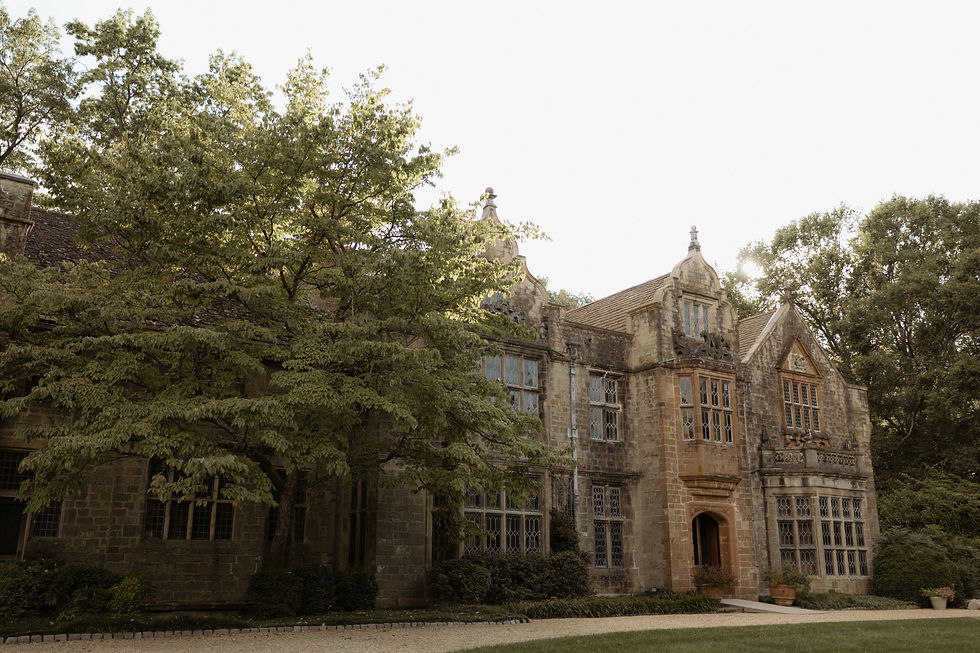 A wide view of the exterior of a historic English Tudor-style stone manor with leaded glass windows and intricate stonework, surrounded by large green trees and a circular gravel driveway.