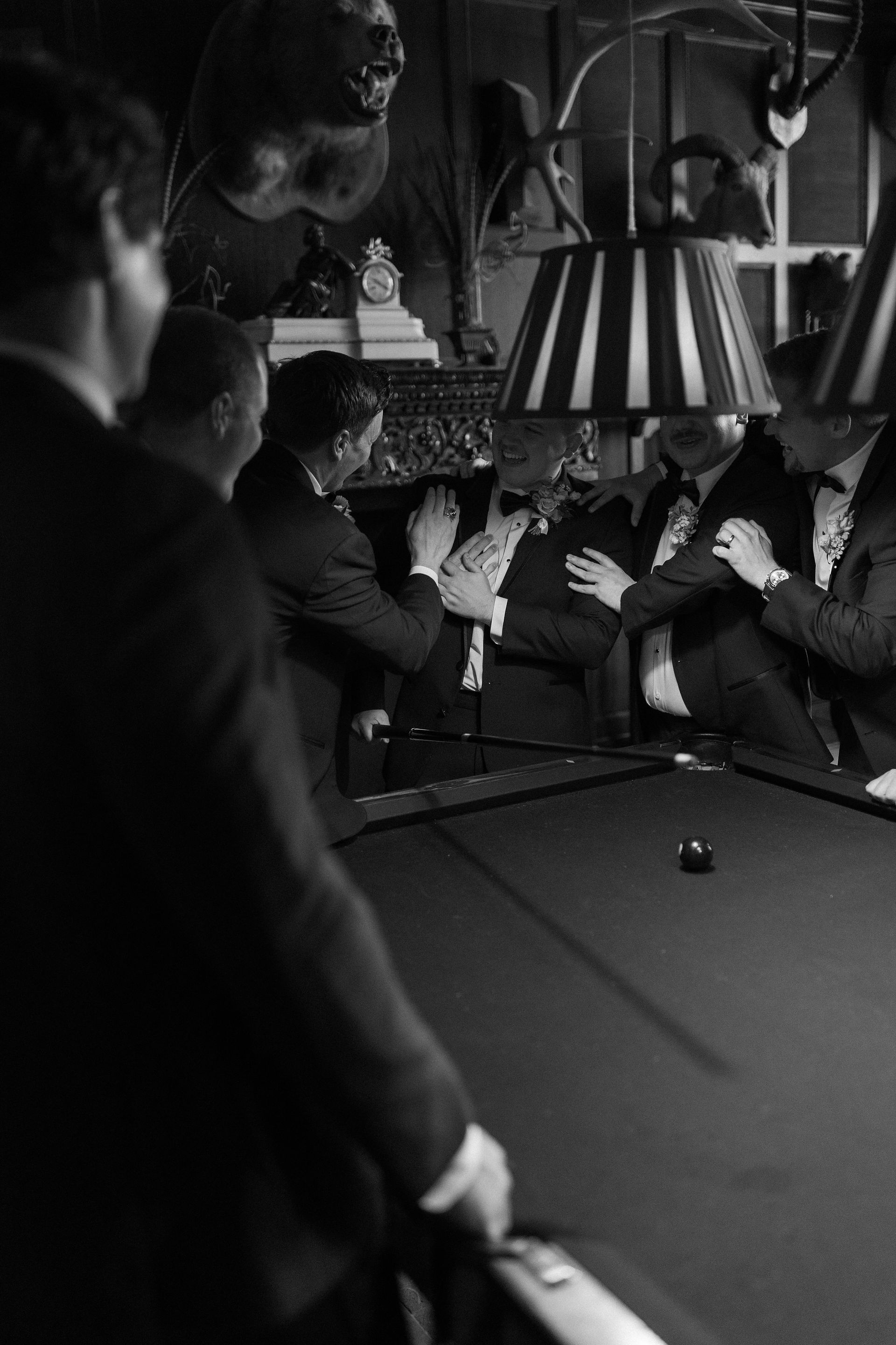 A candid black and white photo of a groom and his groomsmen laughing together around a pool table in a dark, moody billiards room decorated with taxidermy and striped lampshades.