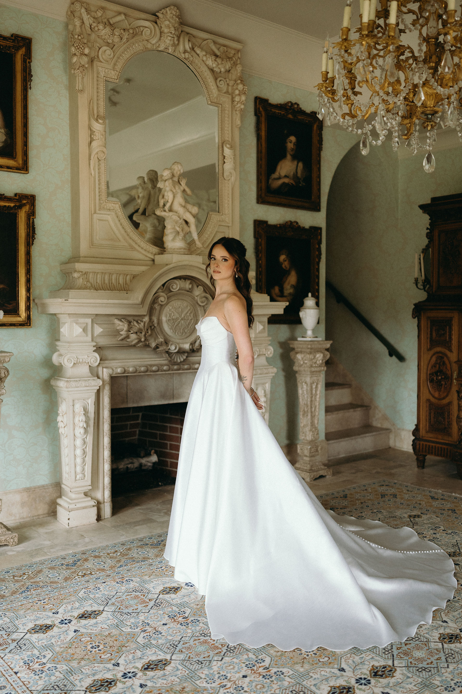 A bride in a classic strapless white wedding gown standing in a lavish room featuring mint green walls, antique oil paintings, a carved fireplace, and an ornate mirror.