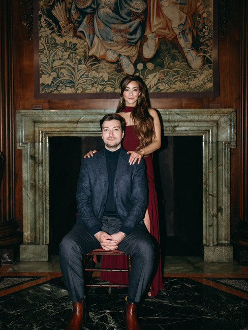 A classic engagement portrait of an elegant couple in a historic, wood-paneled room. The man is seated in front of a grand green marble fireplace wearing a dark blazer, while the woman stands behind him in a burgundy gown, with a large antique tapestry hanging in the background.
