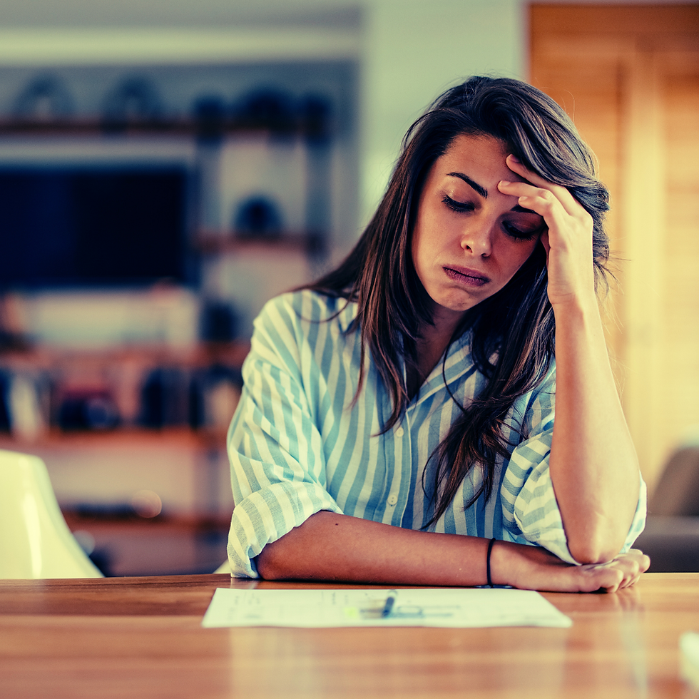 Woman in striped shirt sits at a table, looking stressed with head in hand. Papers are scattered in front of her. Background is blurred.