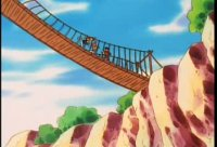 People cross a narrow rope bridge over a rocky canyon under a blue sky, with trees and a bright, adventurous feel.