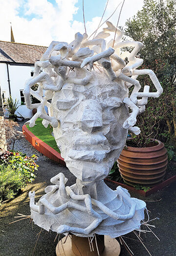 Photograph of work in progress showing a whole front view of the paint finish on the plaster-coated Medusa Head, with the completed ‘snake crown’ attached. The work is taking place outdoors (a garden).