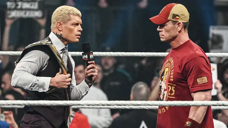 Two wrestlers face off in a ring. One with a microphone and championship belt, wearing a suit. The other in a red shirt and cap. Intense mood.