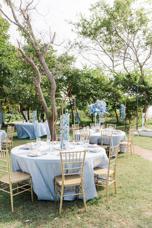 An outdoor wedding reception featuring round tables with blue linens, gold chairs, and floral centerpieces.