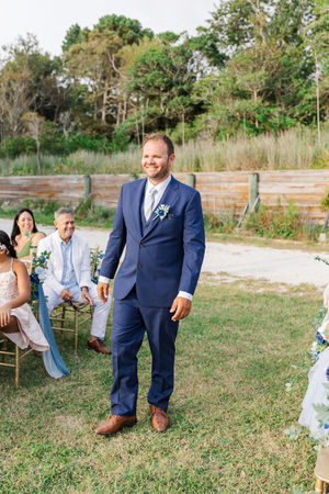 Groom in a navy blue suit walks down the aisle, smiling as he makes his way to the altar at his Virginia Beach wedding.