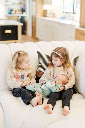 Two older sisters gently holding their newborn baby brother, dressed in a soft green outfit, during an in-home newborn photography session in Richmond, Virginia.