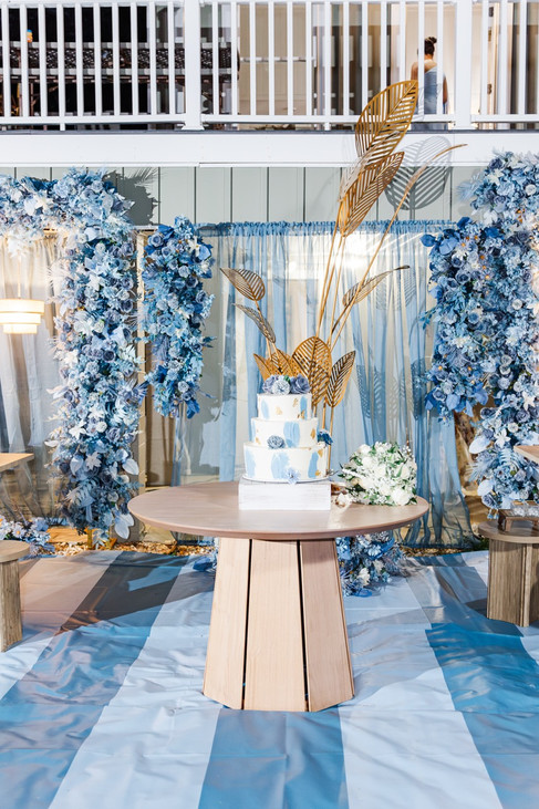 Wedding cake display under a modern floral arch with blue and gold decor accents.