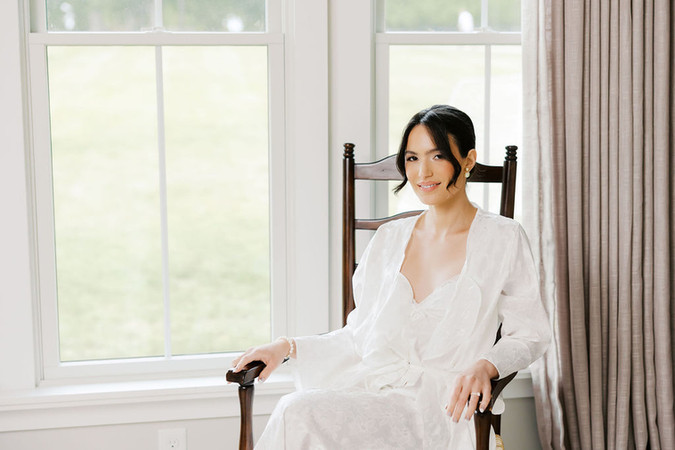 13. Bride sitting by large windows in a white robe during elegant getting ready portraits