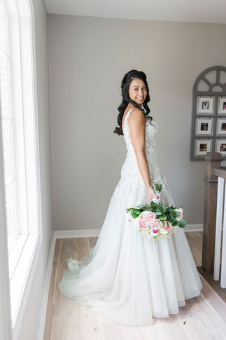 12. Bride standing gracefully, holding her floral bouquet near a window.