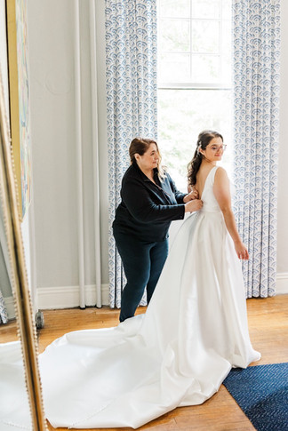 Bride getting ready as a bridesmaid helps fasten her dress in a bridal suite with natural light.