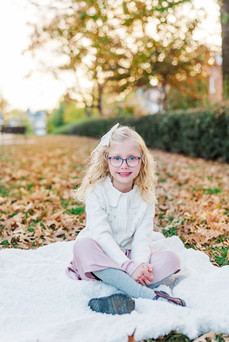 A young girl with blonde curls and glasses sits on a cozy blanket among autumn leaves at the VMFA in Richmond, VA, smiling for her portrait.