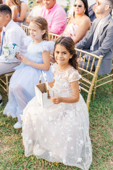 Smiling flower girl in a white lace dress holds a lantern while seated on a gold chair at a coastal wedding ceremony in Virginia Beach.
