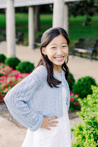 Young girl in a light blue sweater smiling near vibrant garden flowers during a family session.