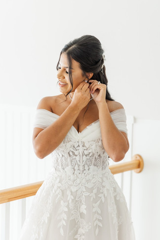 Bride getting ready, smiling as she puts on earrings in an off-the-shoulder lace gown.