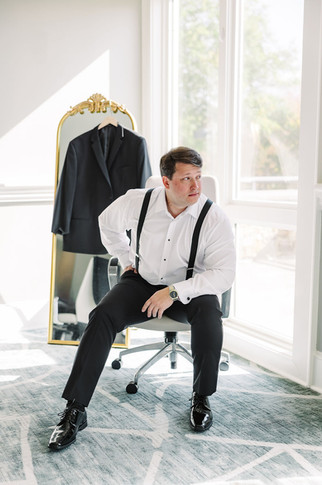 9. Groom seated near window adjusting suspenders — pre-ceremony preparation photo at The Dominion Club.