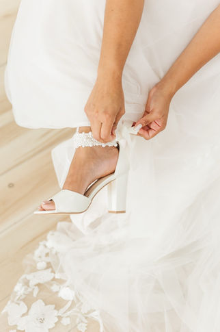 Bride adjusting white block heels during wedding morning preparations.