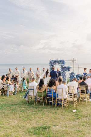 Wedding guests sit on gold chairs, watching the ceremony take place in front of a floral arch with ocean views at a Virginia Beach coastal wedding.