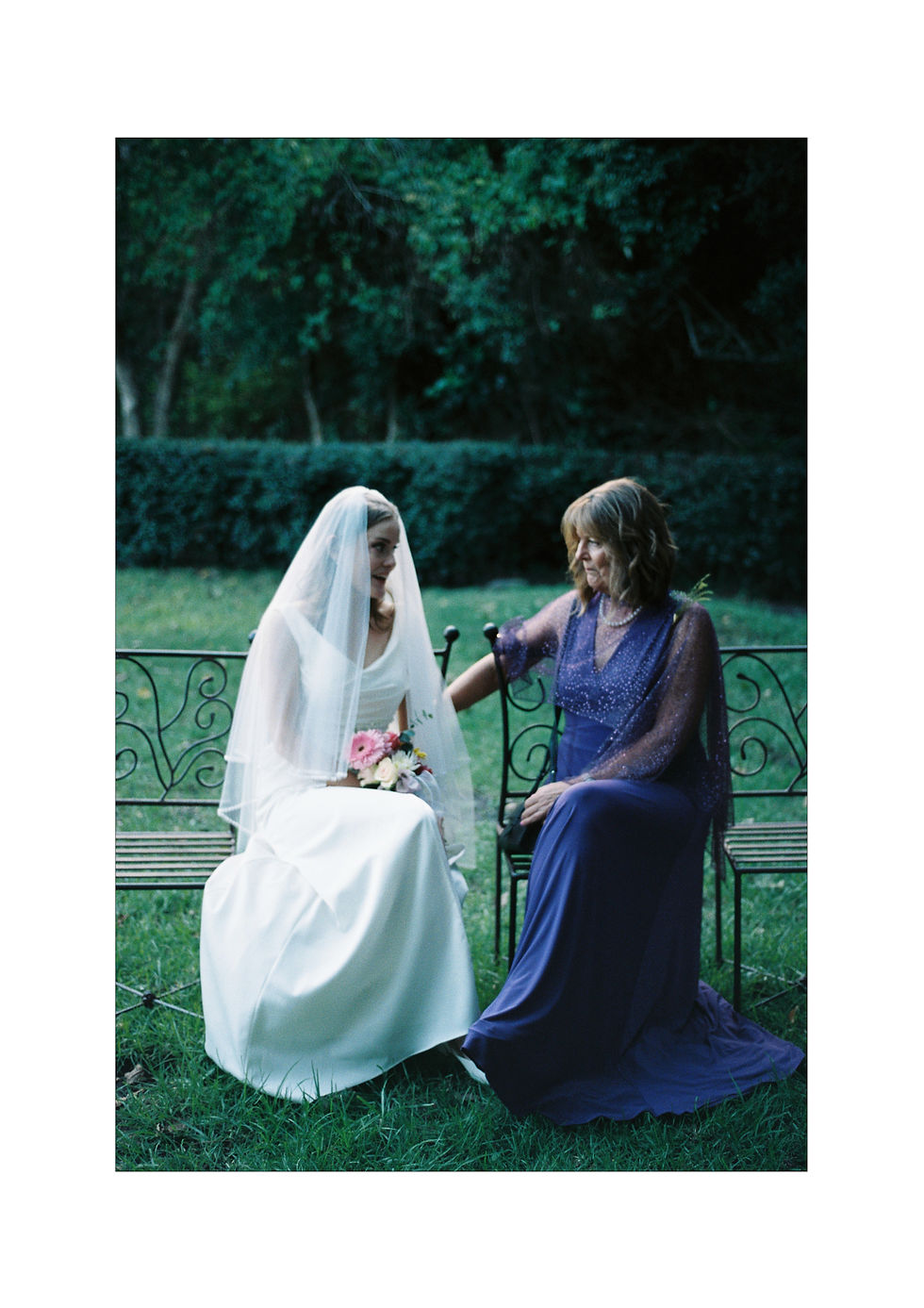 Bride in white seated on bench, veil and bouquet, smiling at woman in purple dress. Greenery in background, serene mood.