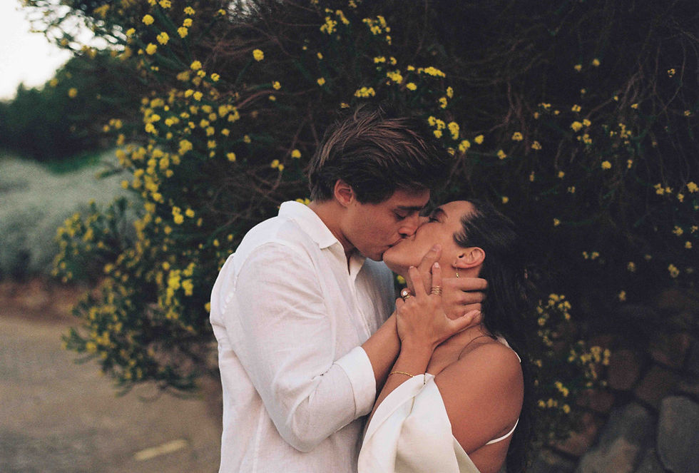 Couple kisses outdoors beside yellow flowers, wearing white, in a romantic close-up.