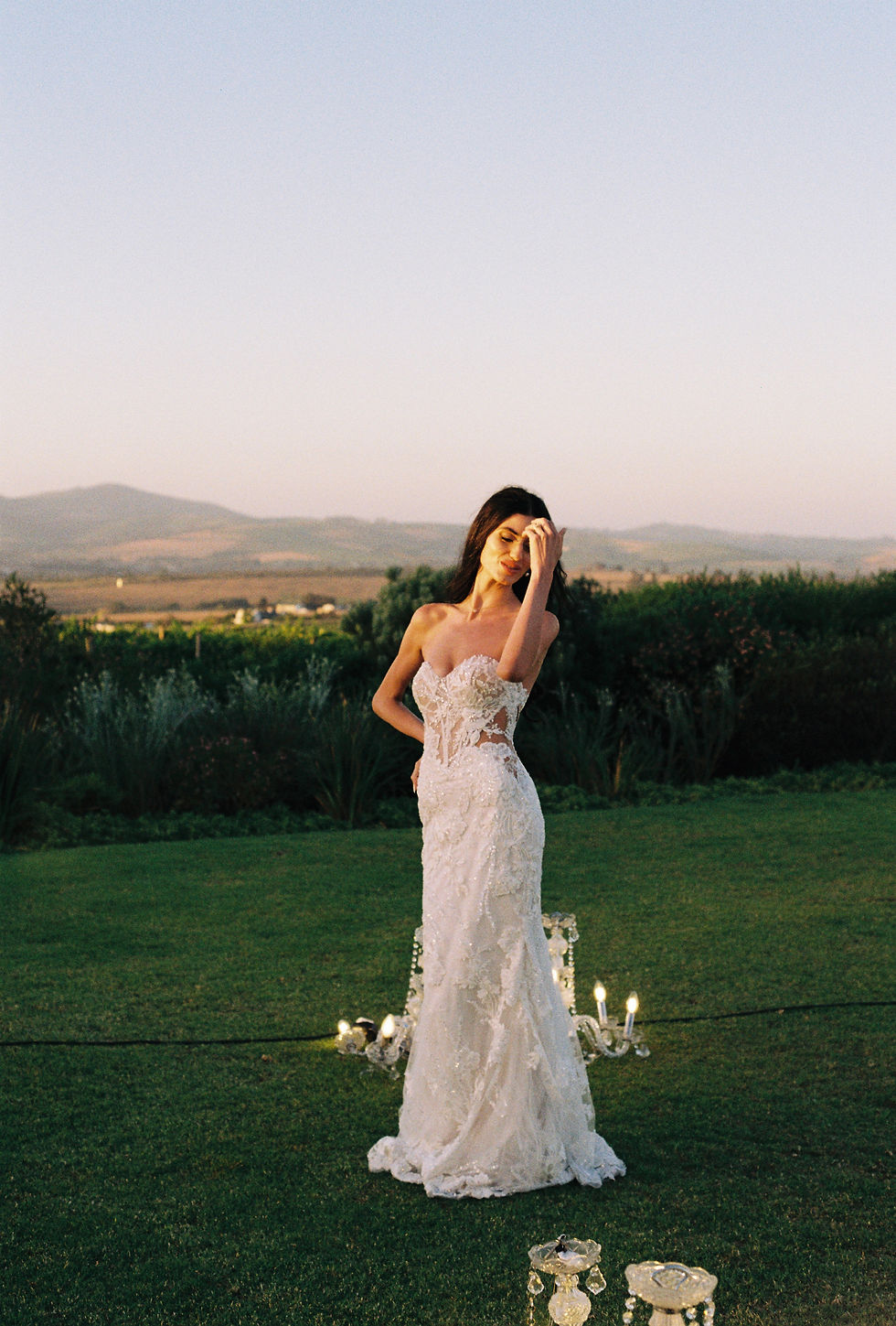 A woman in a white lace gown stands on a grassy field at dusk, surrounded by candles. Mountains and a clear sky form the serene backdrop.