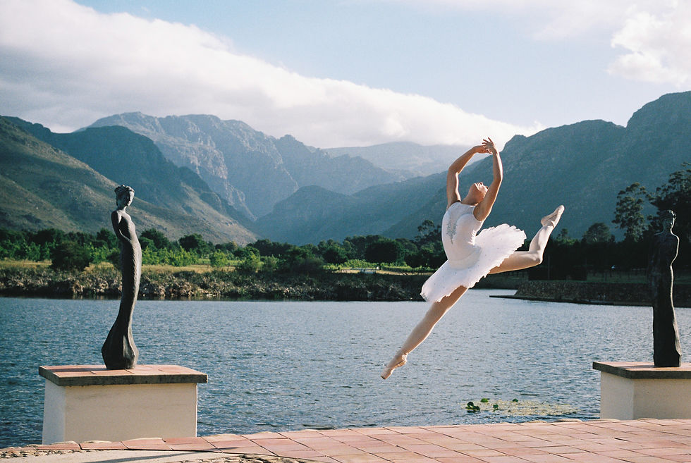 Ballerina in white tutu leaps gracefully by a lake with mountain backdrop. Two statues frame the scene. Sky is partly cloudy.
