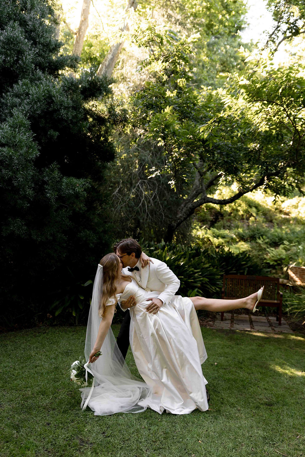 Bride and groom kissing in a lush garden, groom in white tuxedo holds bride in white dress. Sunlit trees and bench in background. Romantic mood.