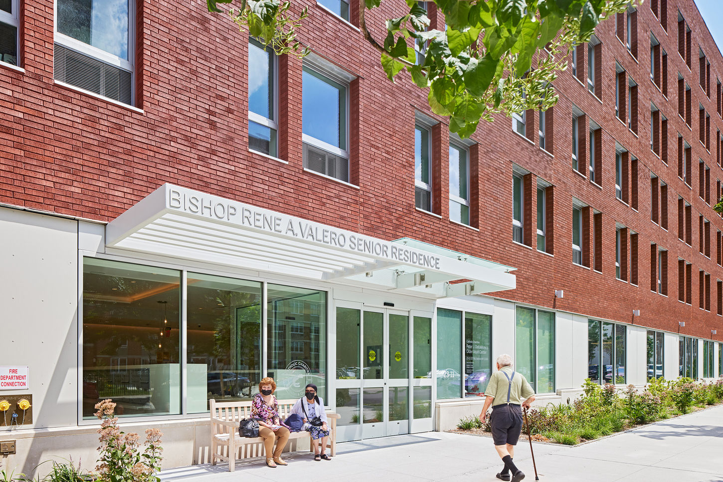 Entrance of the senior housing with two elderly women sitting on the bench and an elder man with a cane walking in.